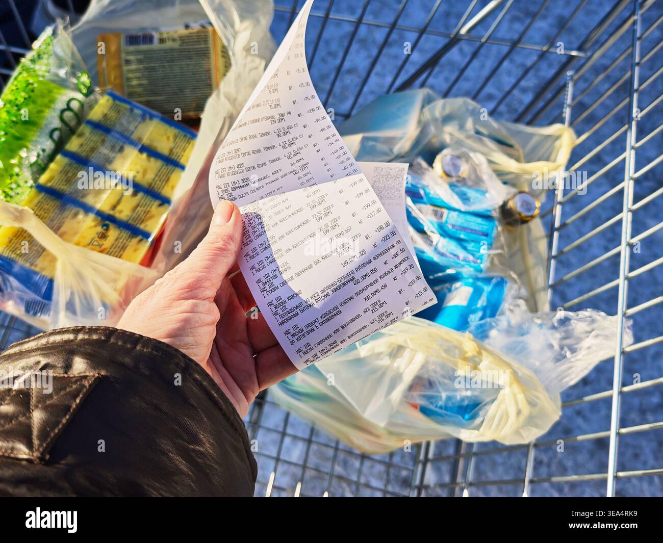 Kirov, Russia - 8 febbraio 2026: Una persona detiene una ricevuta della spesa con un elenco di articoli dopo il check-out al supermercato. Il carrello contiene il sacchetto Foto Stock