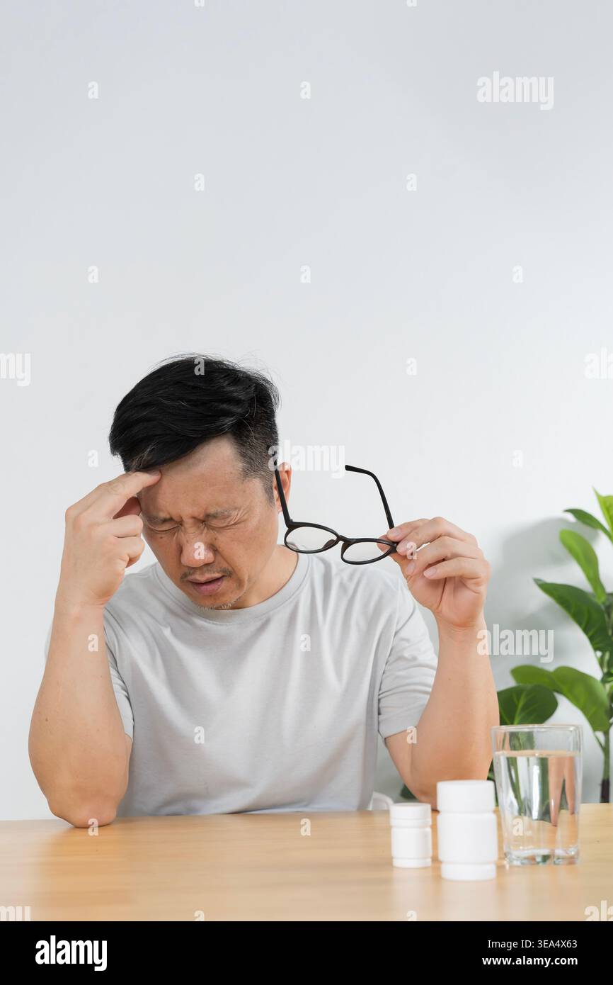 Un uomo asiatico di mezza età preme il tempio con gli occhi chiusi tenendo gli occhiali accanto alla medicina e all'acqua, illustrando mal di testa, affaticamento degli occhi Foto Stock