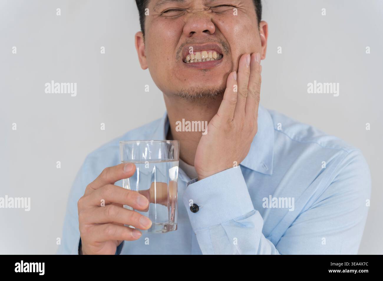 Primo piano di un uomo asiatico che tiene un bicchiere d'acqua mentre preme la guancia nel dolore, illustrando mal di denti, sensibilità dentale, infiammazione orale, e. Foto Stock