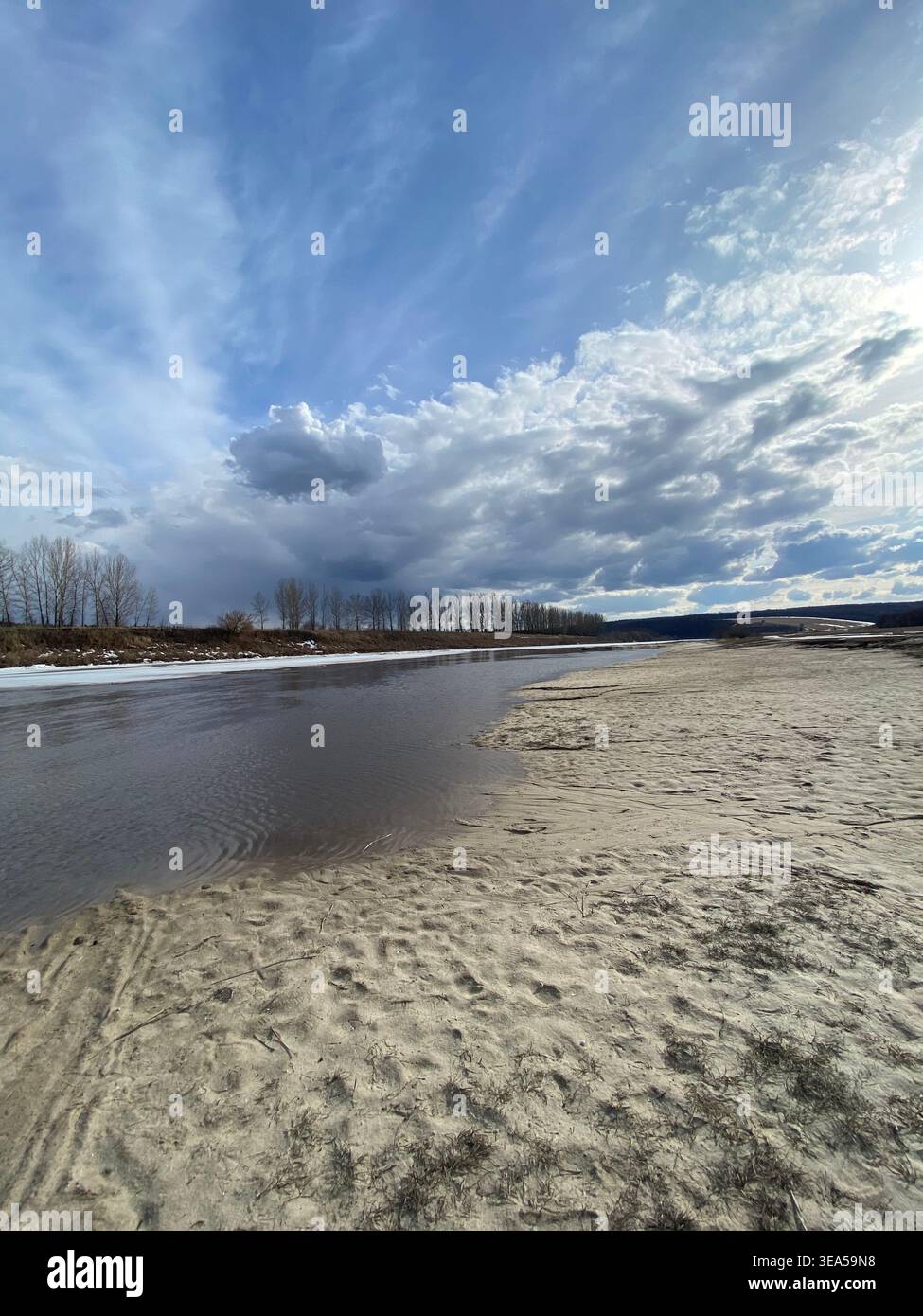 Paesaggio fluviale con spiaggia sabbiosa sotto un cielo nuvoloso, scenario naturale primaverile con acque calme e ampio spazio aperto - Immagine stock catturata con smartphone