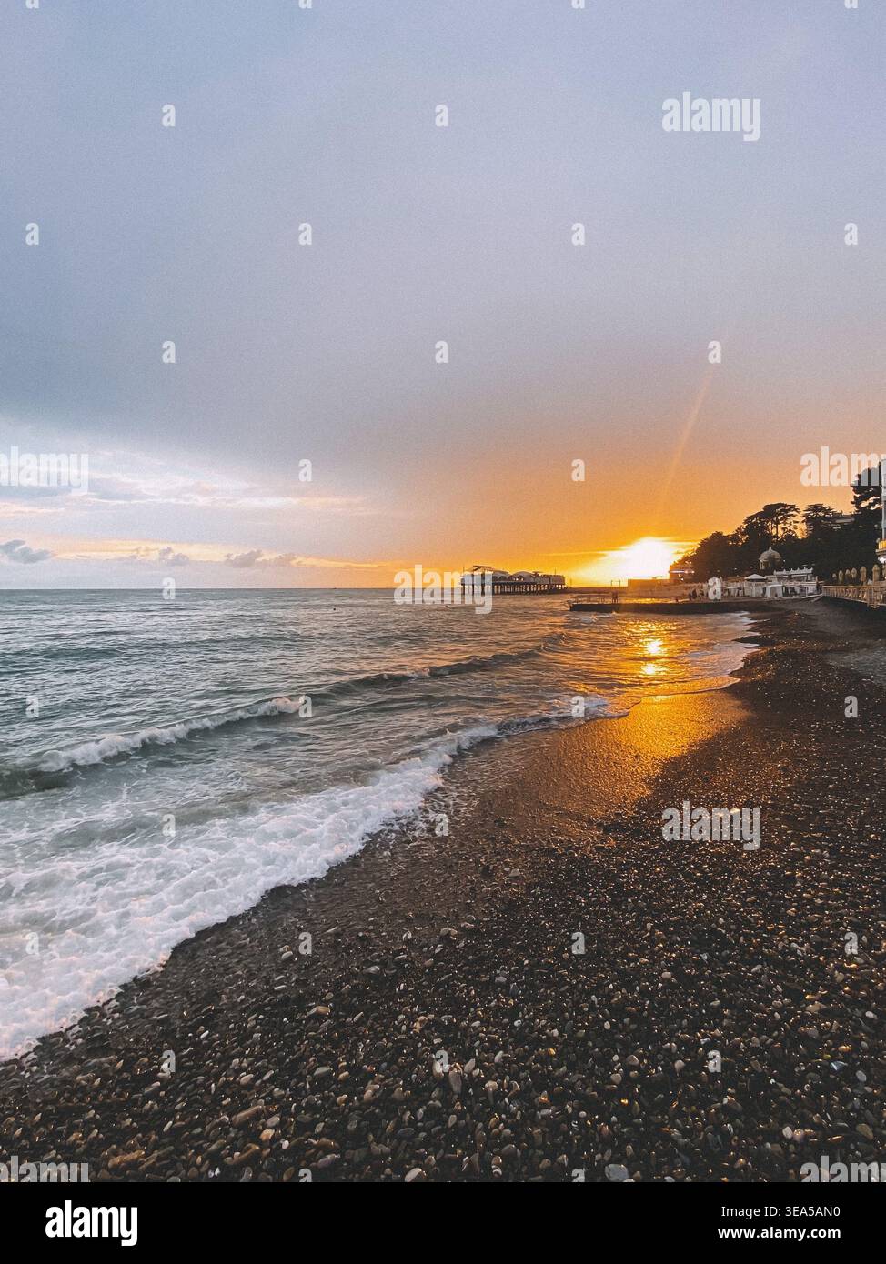 Tramonto dorato sul mare con profilo del molo e onde sulla spiaggia di ciottoli Foto Stock
