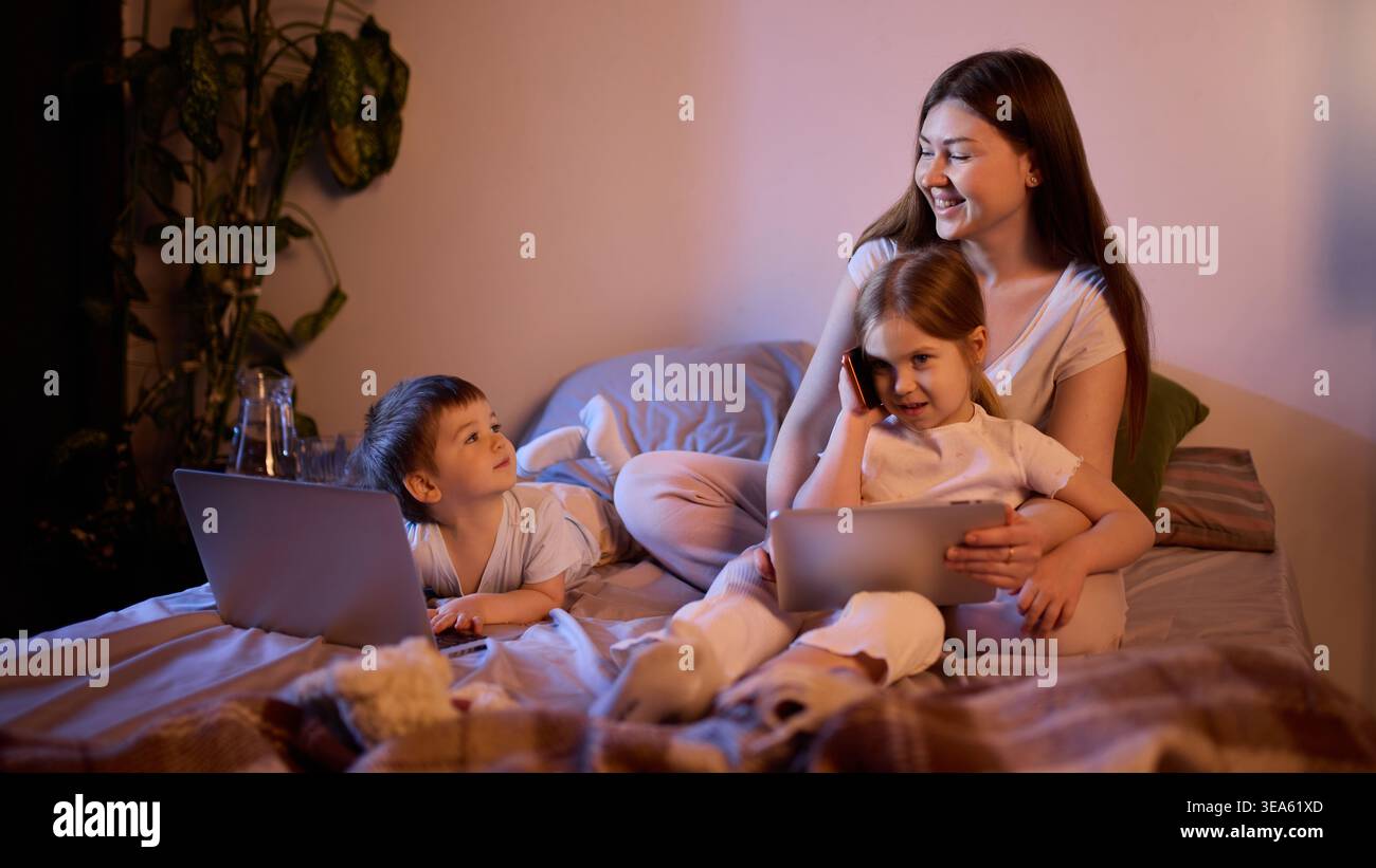 Mamma con bambini che usano tablet e smartphone a letto con una calda luce serale. Foto Stock