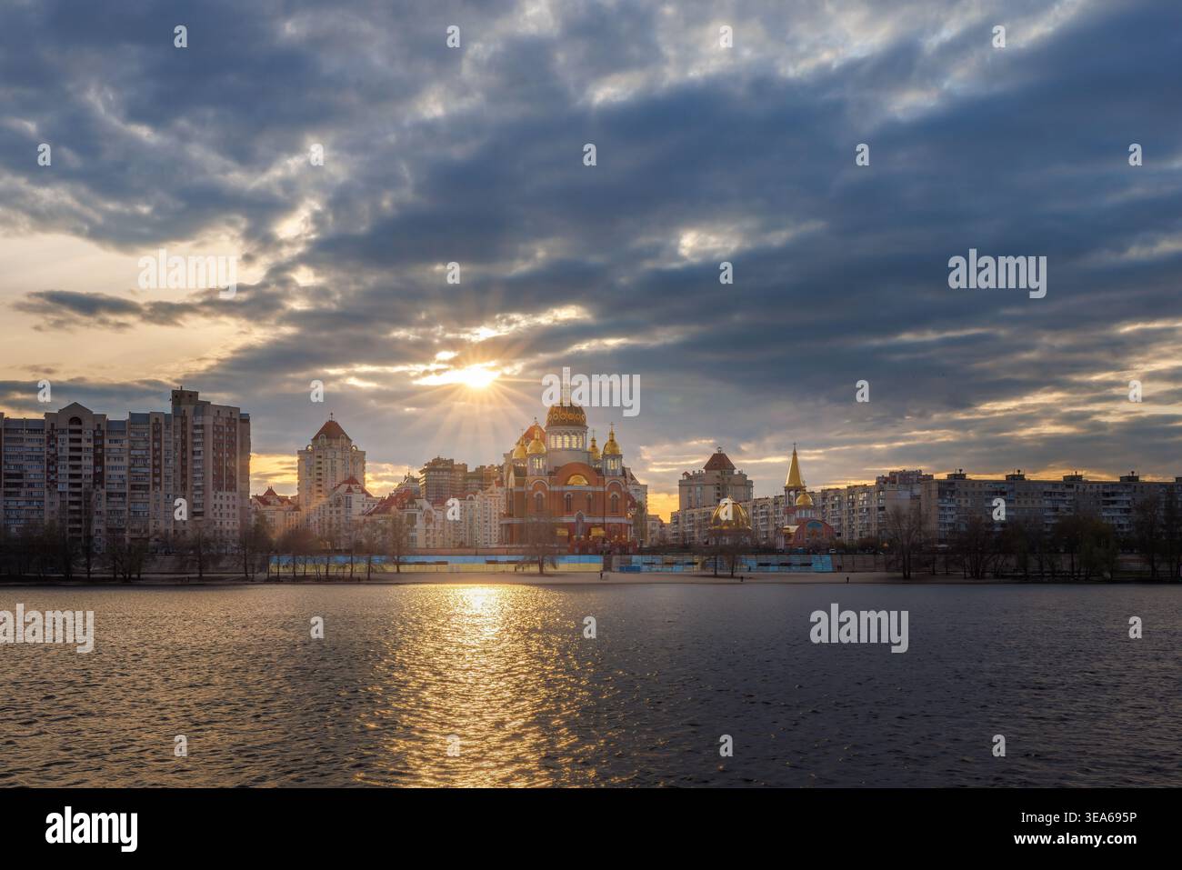 Tramonto dorato sullo skyline di Kiev sull'argine del fiume Dnipro, caldi raggi del sole che si riflettono sull'acqua, spettacolari nuvole e architettura urbana Foto Stock