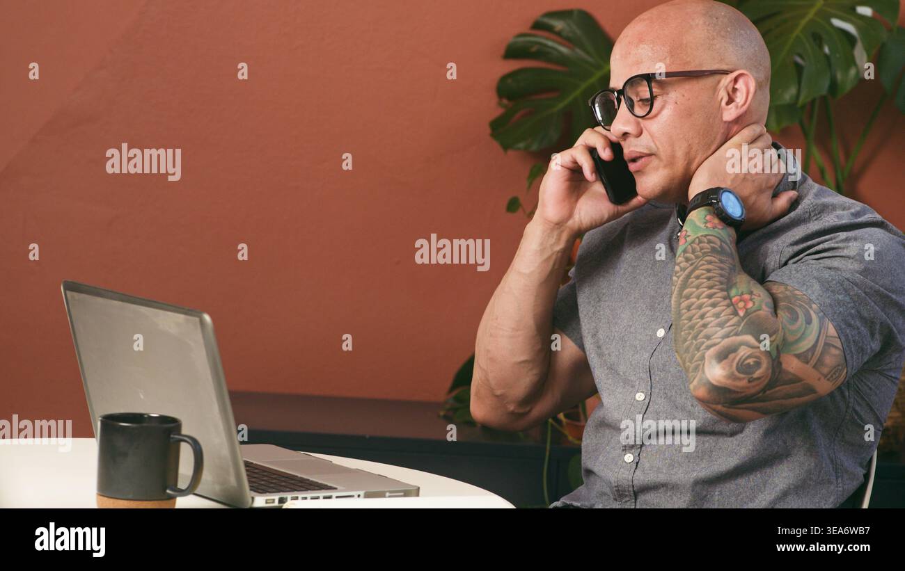 Uomo d'affari tatuato che effettua una telefonata mentre lavora su un laptop in un moderno ufficio domestico Foto Stock