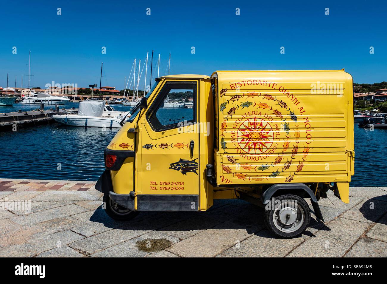 Un piaggio Ape giallo a tre ruote per il ristorante Gianni al Molo parcheggiato presso il porto turistico di Porto Rotondo in Sardegna, Italia, con yacht sullo sfondo. Foto Stock
