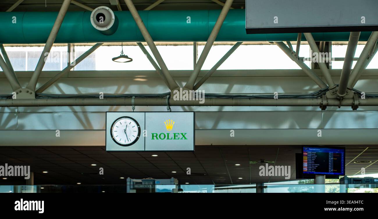 Orologio da parete Rolex e logo all'aeroporto di Olbia Costa Smeralda, Italia. L'orologio analogico è appeso alla struttura a soffitto industriale del terminale. Foto Stock