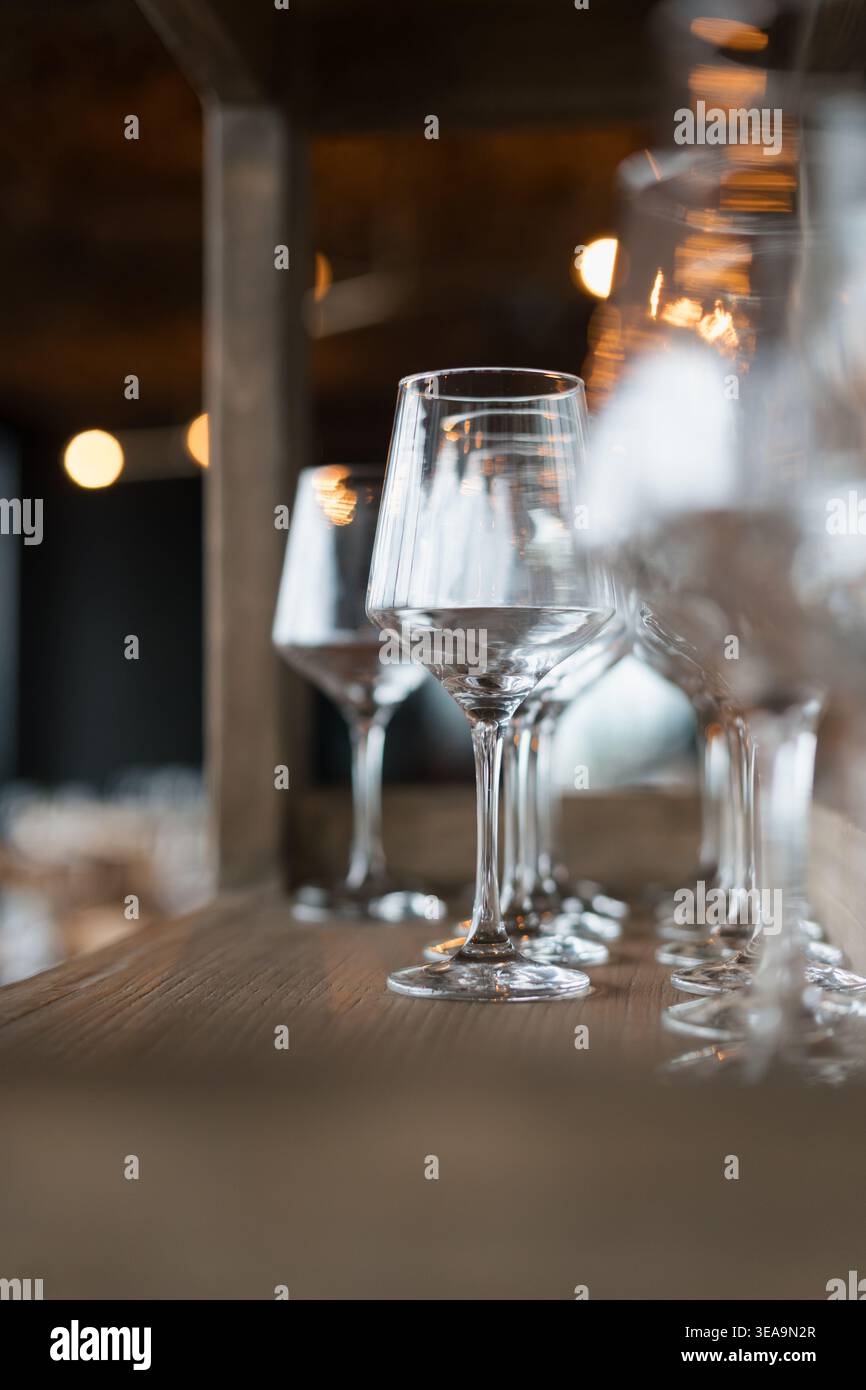 I bicchieri da vino trasparenti si trovano in fila su un tavolo di legno. Le luci soffuse sfocano lo sfondo. La messa a fuoco è nitida sugli occhiali in primo piano. Un'atmosfera calda lo suggerisce Foto Stock