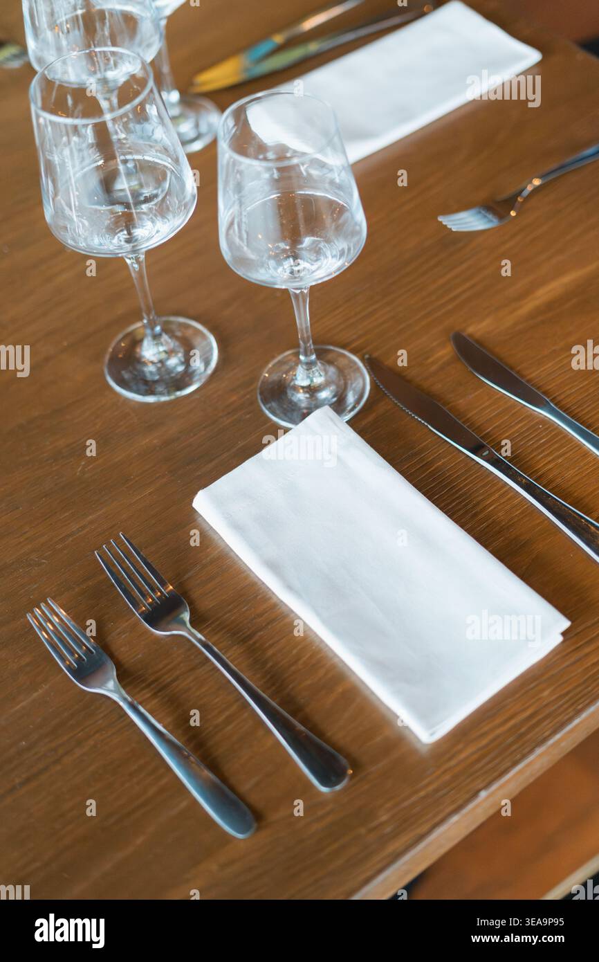 Il tavolo in legno regge un elegante allestimento da pranzo. I bicchieri da vino trasparenti rimangono vuoti sulla superficie. Due forcelle e coltelli poggiano ordinatamente accanto al tovagliolo. Nappa bianca Foto Stock