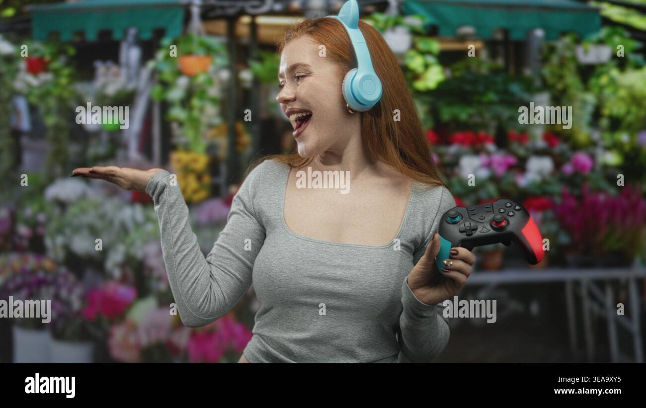 Donna rossa che tiene in mano il gamepad e indossa cuffie con orecchie di gatto, gesti palmari in un mercato dei fiori di strada; gioia giocosa. Foto Stock