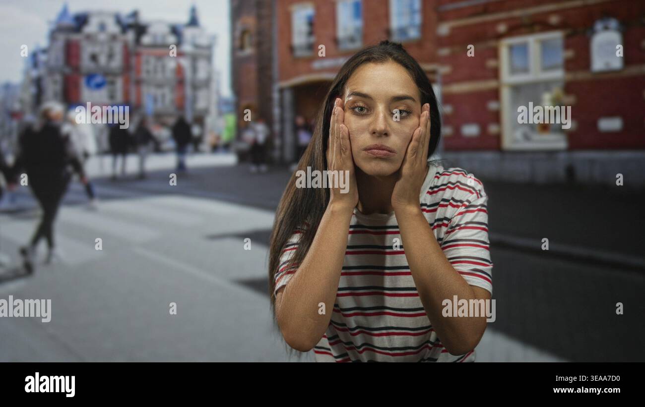 Una giovane donna tiene i capelli tra le dita e ispeziona le radici mentre si trova su un attraversamento di strade trafficate della città vicino a edifici in mattoni che indossano un An bianco a righe Foto Stock