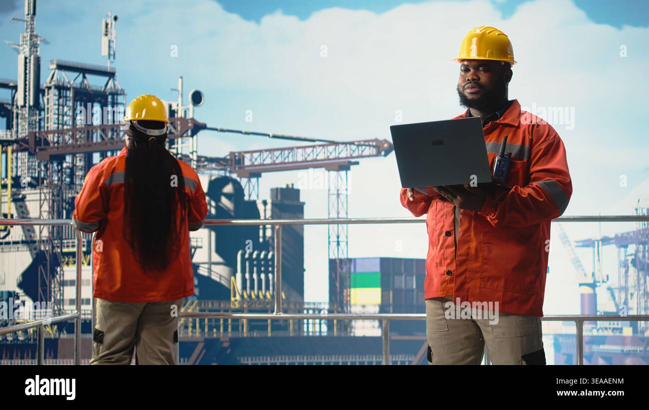 Uomo che lavora per le operazioni di monitoraggio di piattaforme offshore industriali sul ponte utilizzando un computer portatile. Dipendente di un carro di perforazione afroamericano che esamina le operazioni energetiche utilizzando un notebook, Foto Stock