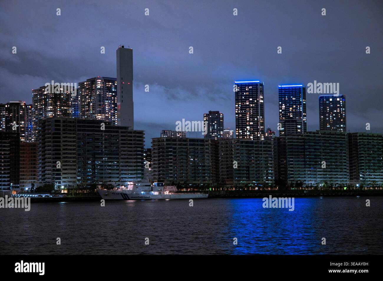 Le moderne e alte torri residenziali illuminate da accenti LED blu si innalzano sul buio lungomare di Tokyo di notte, una grande nave ormeggiata sotto. Paesaggio notturno urbano moodico. Foto Stock
