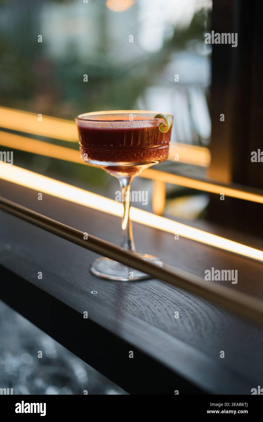 Il cocktail scuro poggia su un davanzale scuro. Riempie un vetro a gambo di una ricca tonalità ambra. La modanatura verde adorna il bordo. Le strisce luminose calde illuminano così Foto Stock