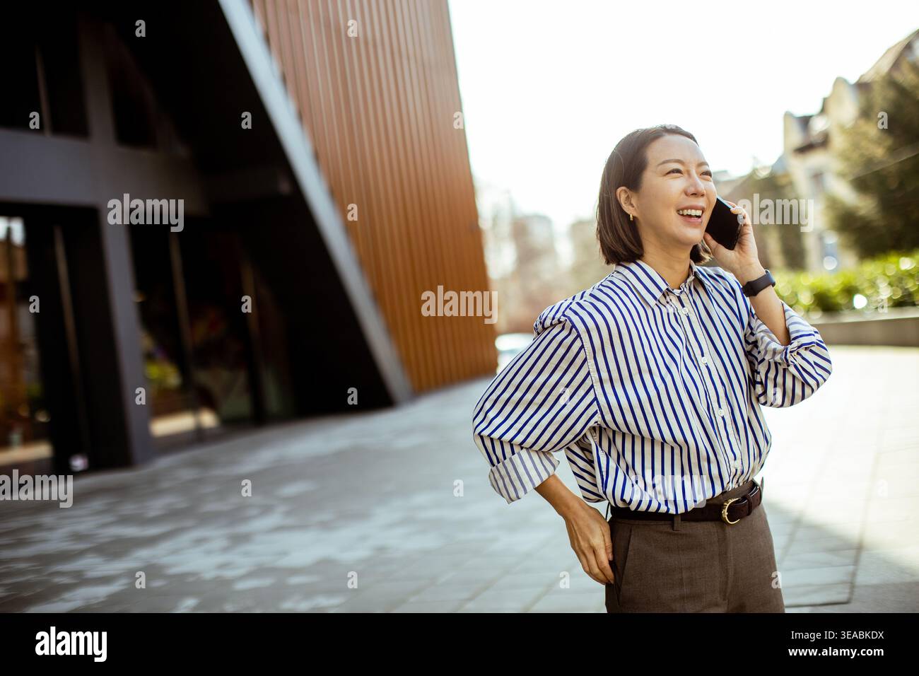 Una donna coreana si trova fuori da un edificio moderno. Tiene un telefono cellulare all'orecchio e sorride mentre parla. Il sole splende su di lei e sul pa Foto Stock