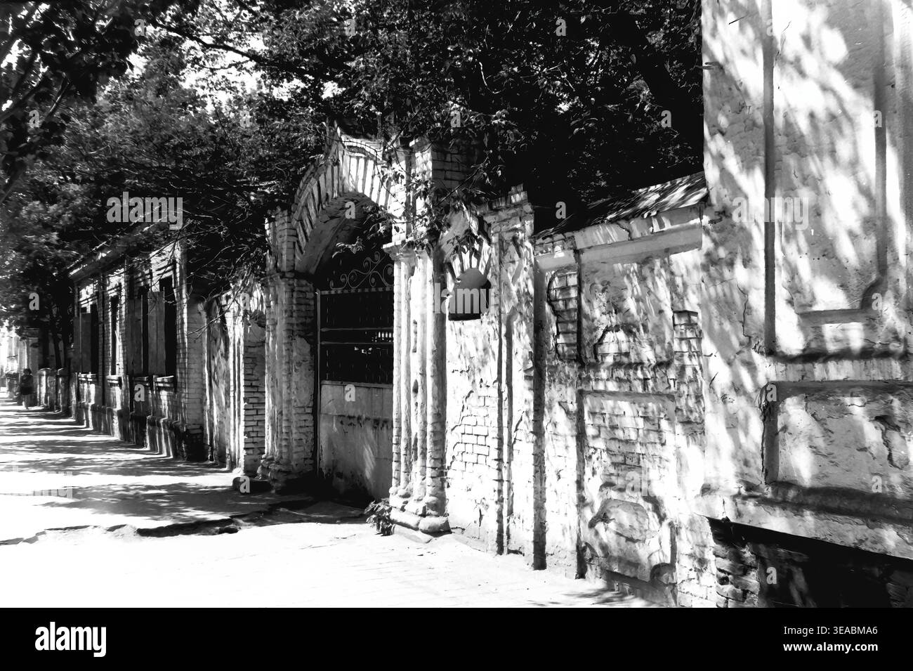 Slavyansk, 1970 anni Vista dettagliata a livello della strada che si concentra direttamente su un arco storico in mattoni e una spessa parete di confine vicino ai grandi magazzini in Karl Marx Street (ora Tsentralna Street). L'intensa luce del sole proietta ombre nette dal fogliame circostante, enfatizzando l'intonaco sbriciolato e il decadimento architettonico. Questa accattivante fotografia d'archivio in bianco e nero funge da monumentale testimonianza del patrimonio urbano della città anteguerra, immortalando splendidamente le tranquille e senza tempo strade del tranquillo Donbas. Foto Stock