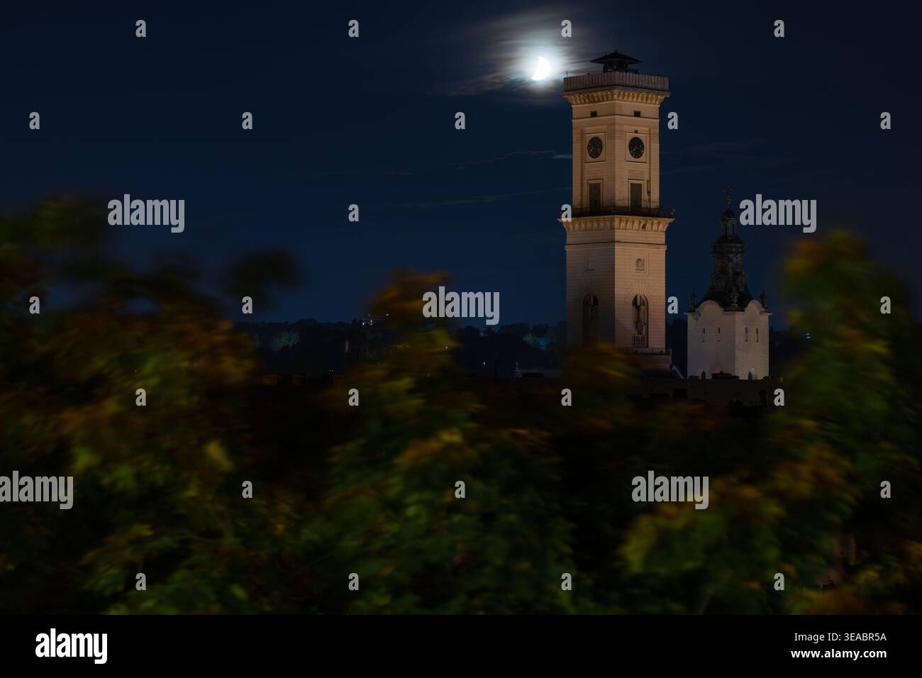 Una storica torre dell'orologio del municipio europeo è illuminata contro il cielo notturno con una luna crescente visibile attraverso nuvole di neve Foto Stock