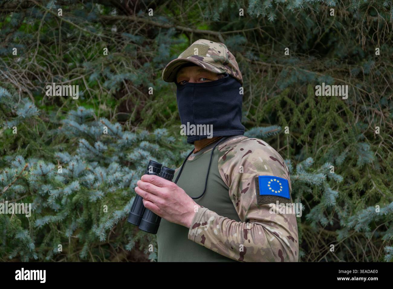 Un soldato con una patch dell'Unione europea conduce la sorveglianza attraverso un binocolo e controlla la situazione sul campo Foto Stock