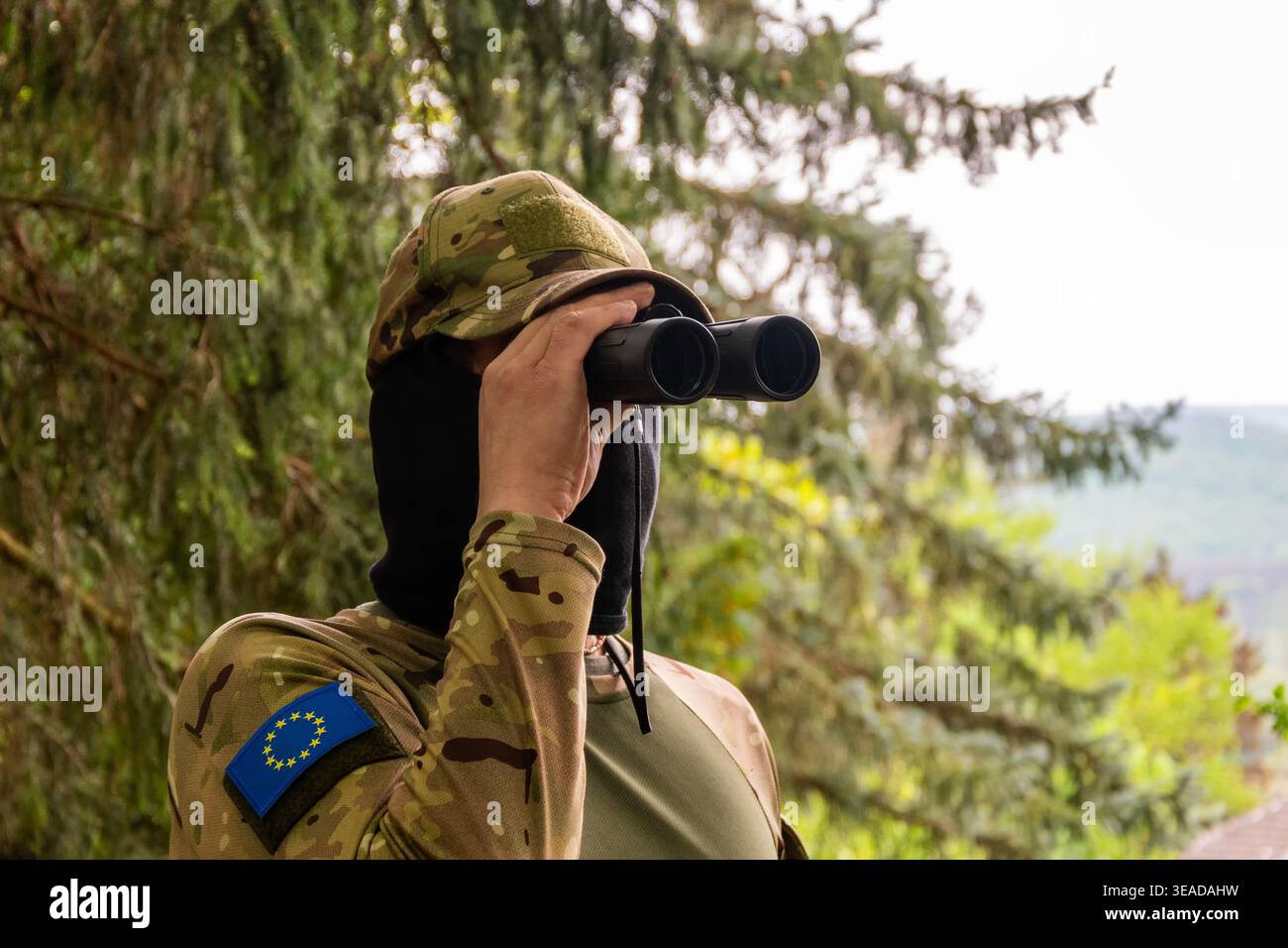 Un soldato con una zona dell'Unione europea osserva attraverso un binocolo e monitora la situazione in un territorio lontano tra foreste e colline Foto Stock