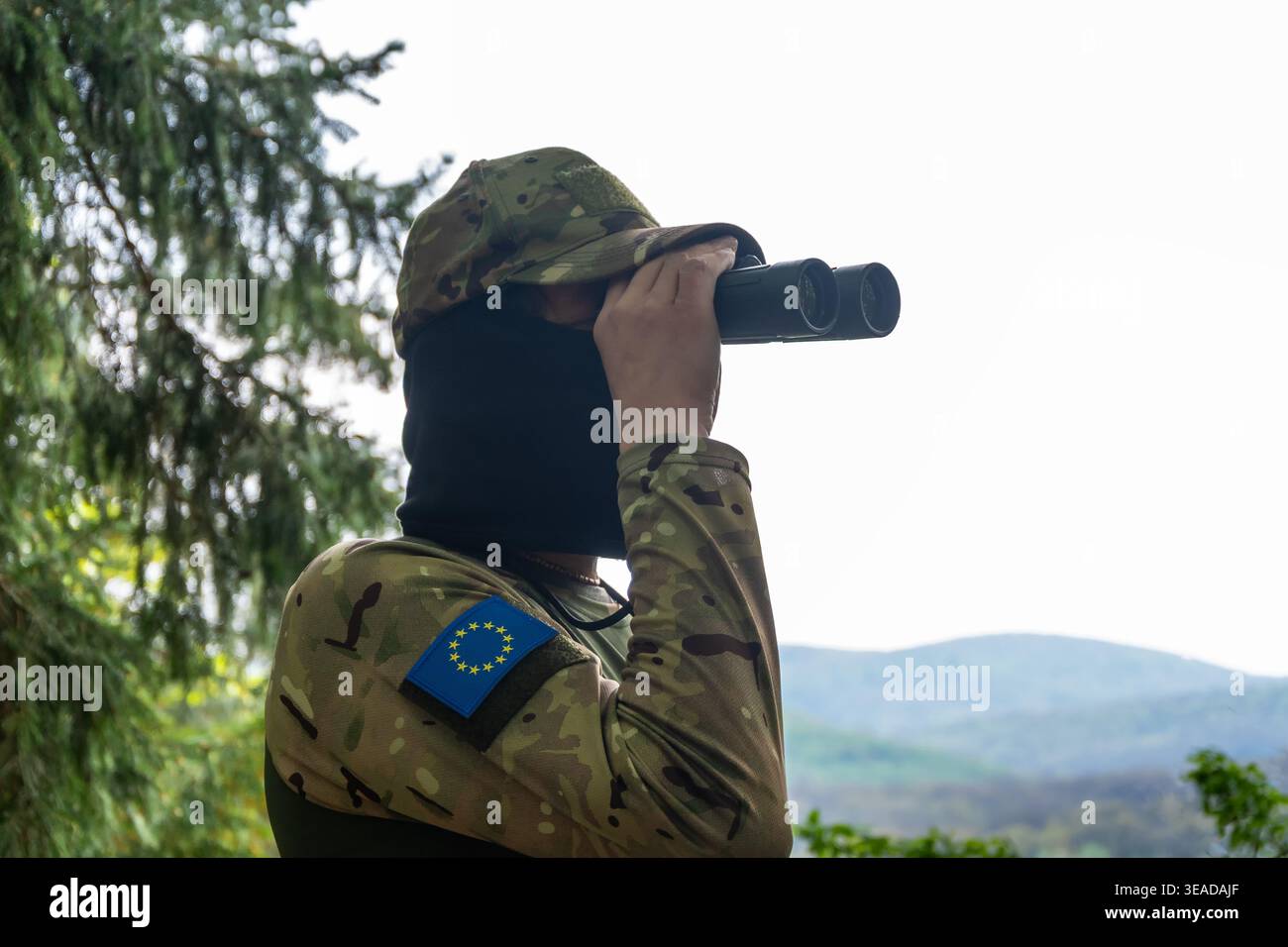 Un soldato con una zona dell'Unione europea conduce la sorveglianza attraverso un binocolo e monitora il confine in un'area boschiva di montagna Foto Stock