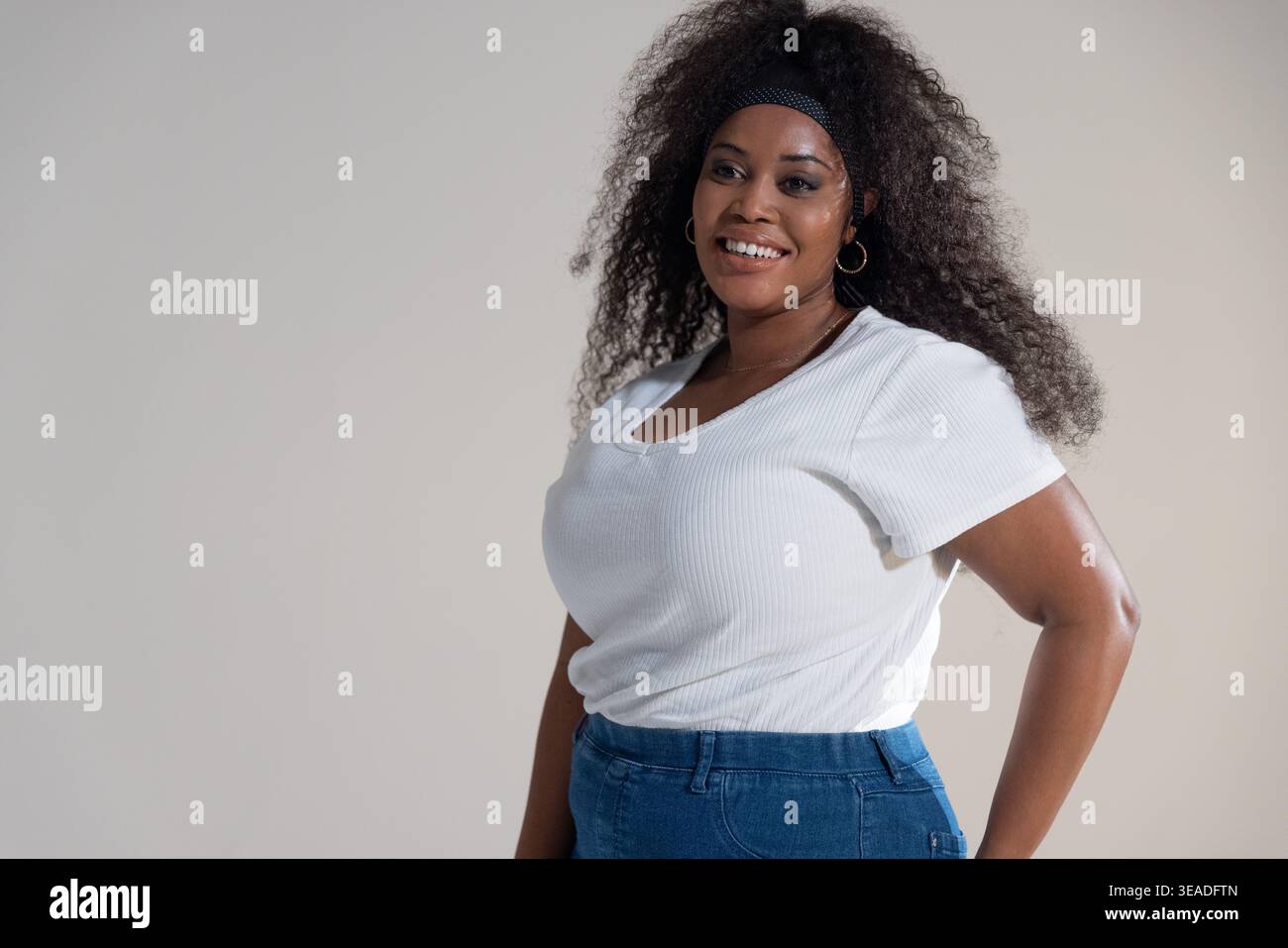 Donna afroamericana di metà età che posa in studio indossando jeans top bianchi fascia nera con cerchi dorati Foto Stock