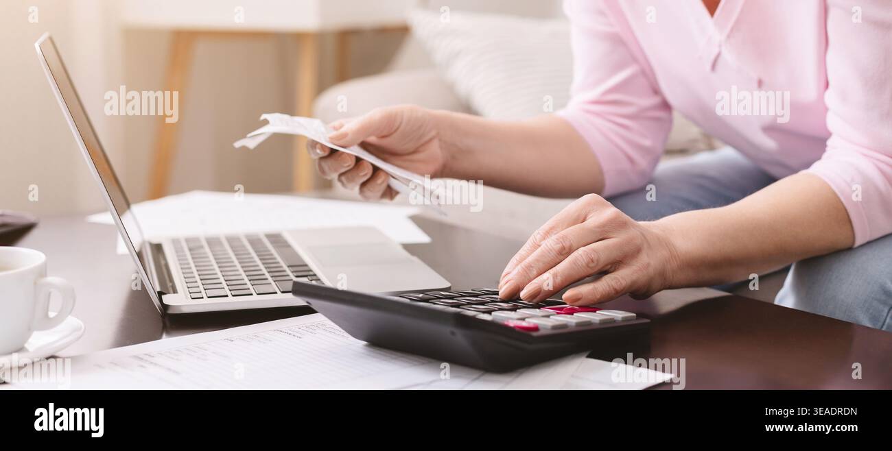 La persona lavora a casa utilizzando la calcolatrice e il laptop con i documenti sul tavolo mentre tiene in mano le ricevute Foto Stock