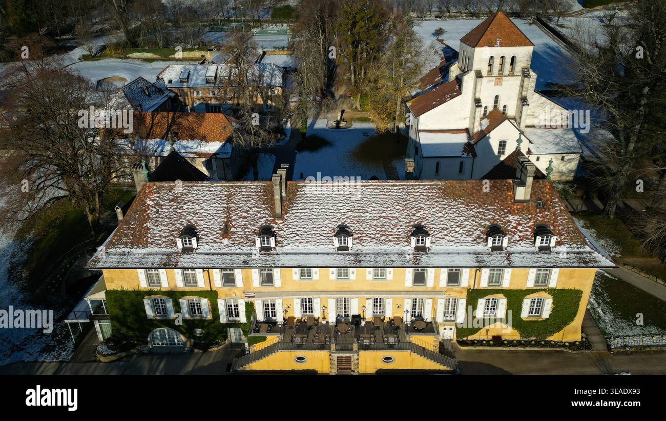 Veduta aerea di un grande edificio giallo con un tetto di tegole rosse, contrastato dai terreni innevati e dalla torre della chiesa adiacente, Chéserex, Vaud, Svizzera Foto Stock