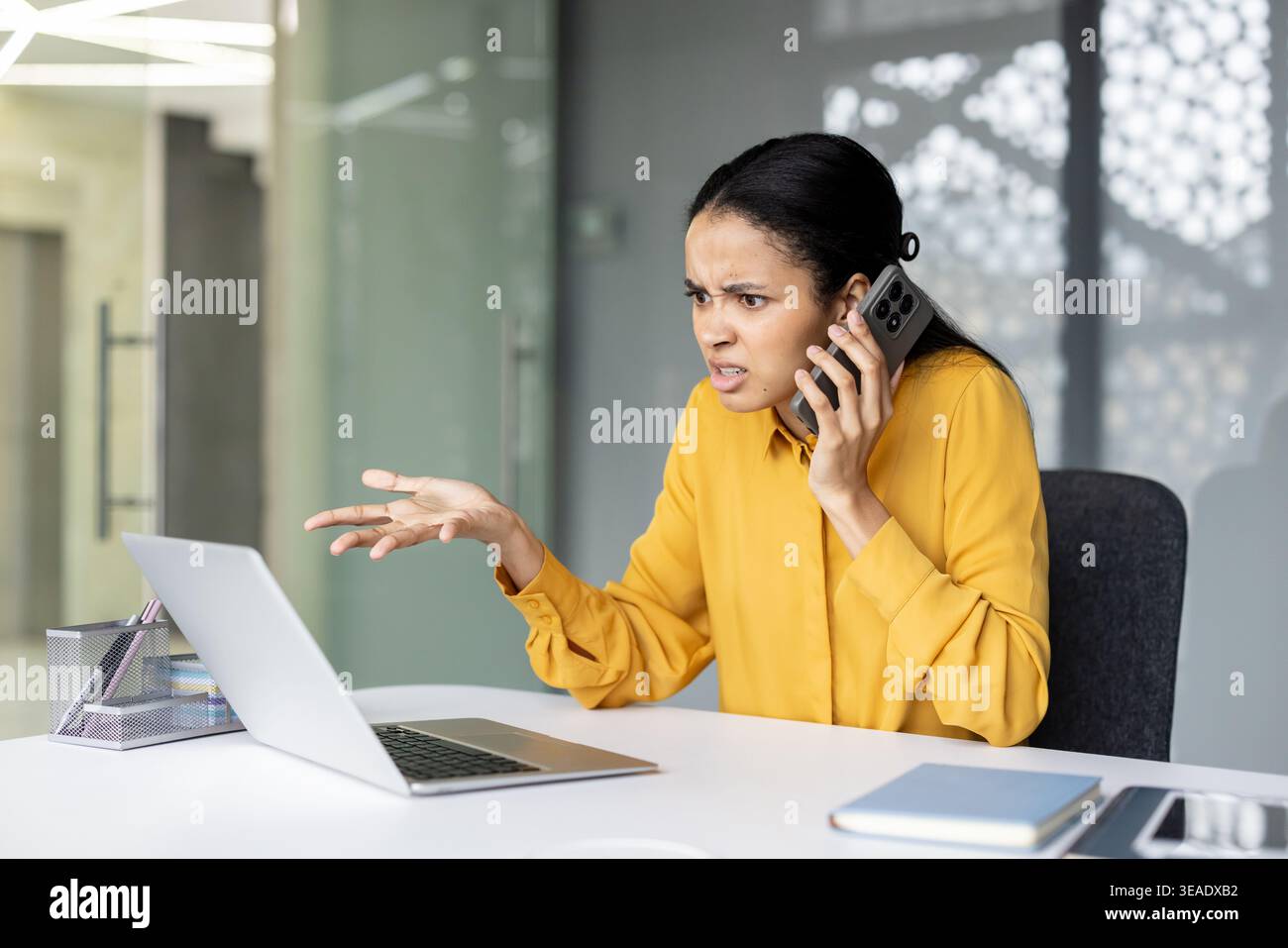 Una giovane donna d'affari frustrata che ha una conversazione arrabbiata al telefono cellulare, utilizza un notebook, sperimenta stress e problemi durante il lavoro in un ufficio moderno Foto Stock