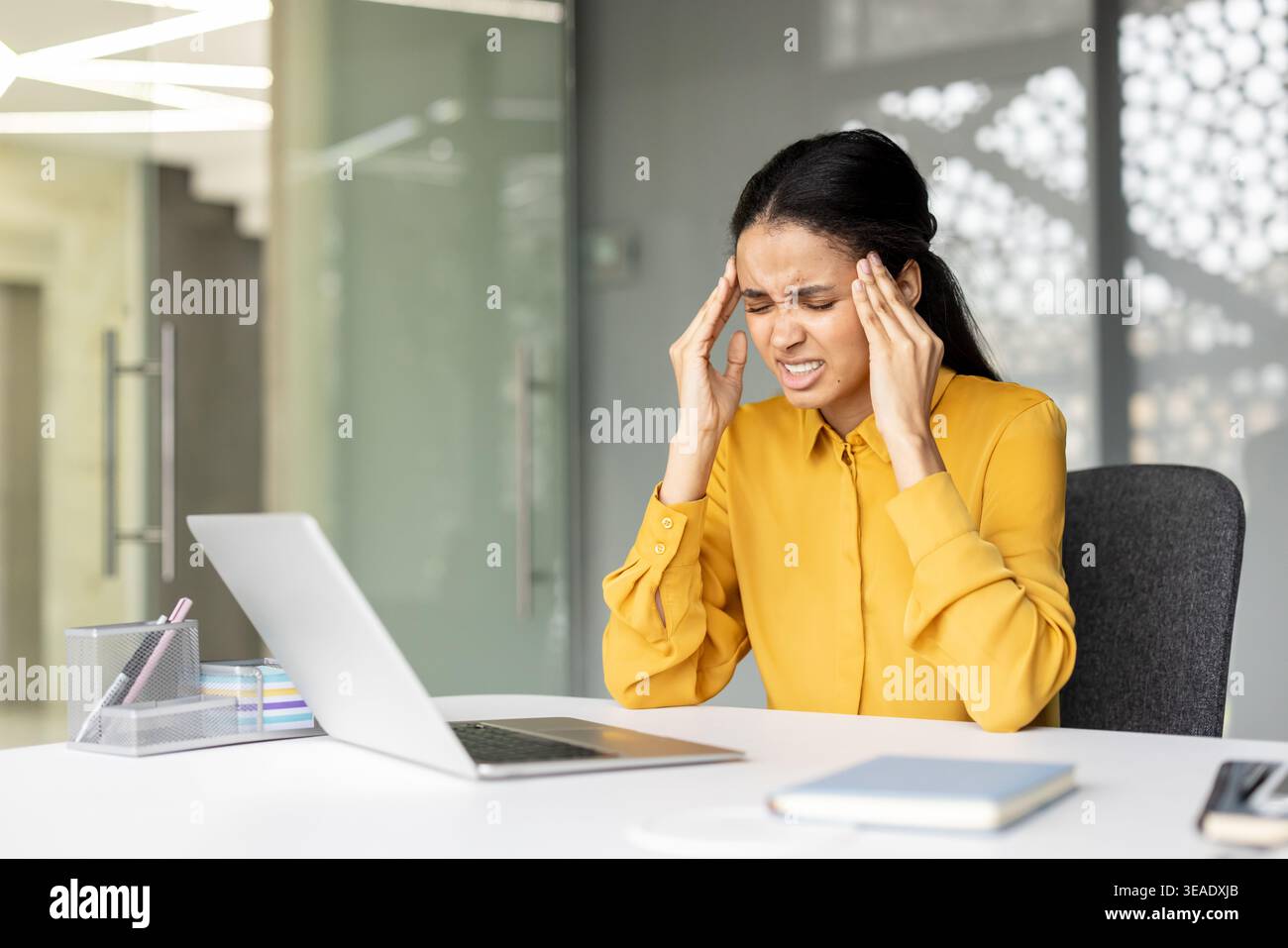 Una giovane donna stressata che lavorava alla sua scrivania sviluppava un forte mal di testa, pressava le tempie con le mani, mentre sperimentava dolore, burnout ed affaticamento emotivo dovuto alla costante pressione sul lavoro Foto Stock