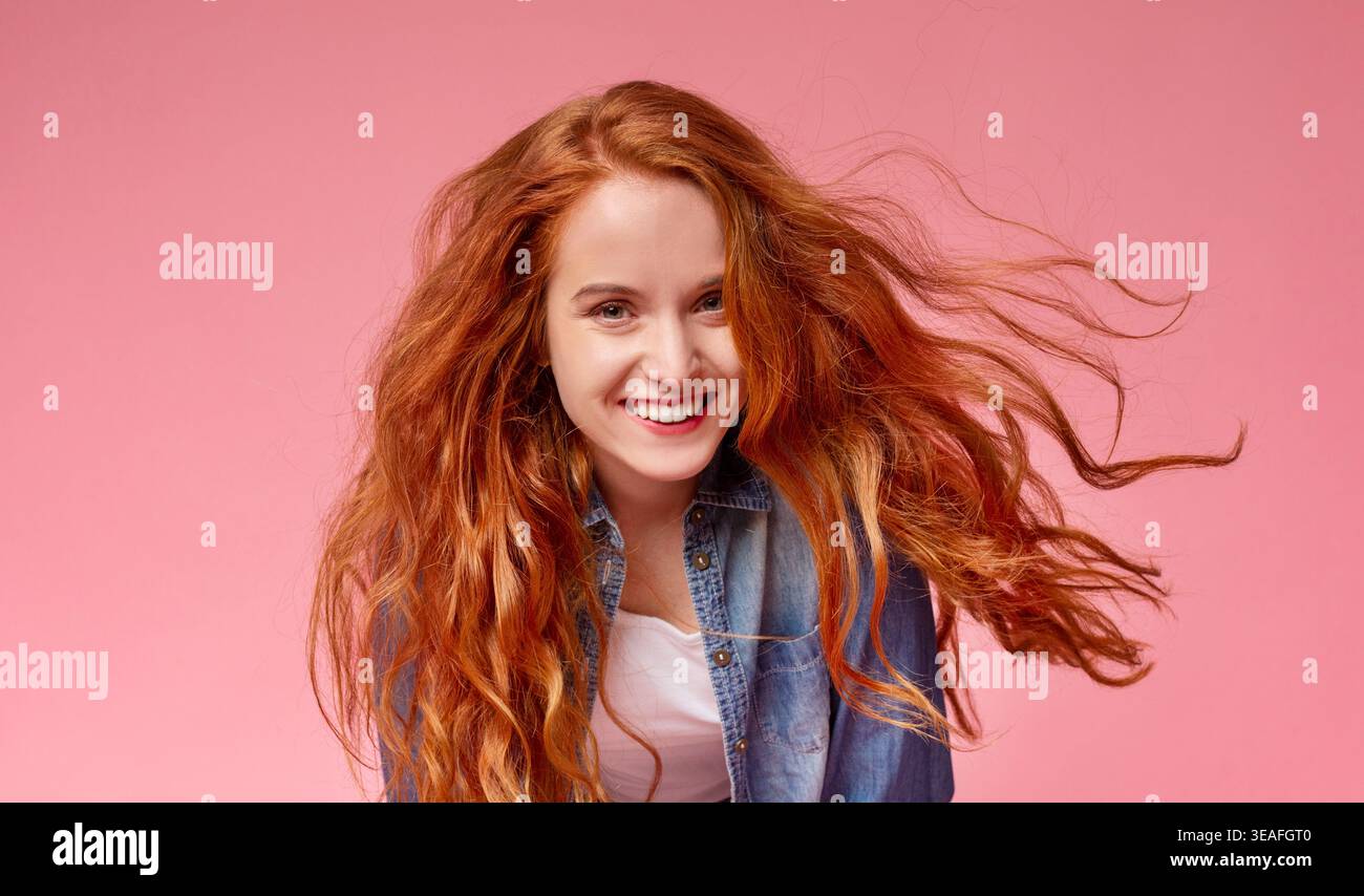 La giovane donna con lunghi capelli rossi sorride gioiosamente su uno sfondo rosa mentre posa giocosamente Foto Stock