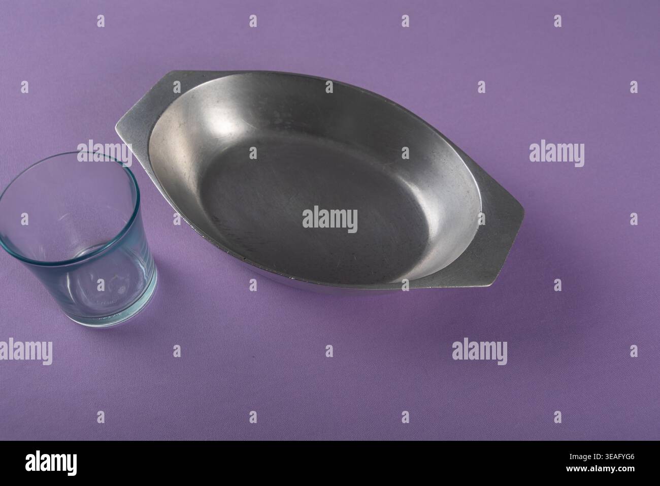 Natura morta minimalista su sfondo viola. Vassoio da forno argento e vetro blu. L'illuminazione soffusa e diffusa mette in risalto le trame metalliche e di vetro. Foto Stock