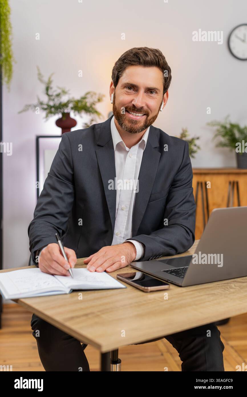 Un uomo d'affari di mezza età in ufficio a casa guarda il webinar sul laptop che indossa auricolari prende appunti e sorride. Il freelance al tavolo scatta la foto dello schermo dello smartphone e tocca il tasto per cambiare la diapositiva oggi stesso. Foto Stock