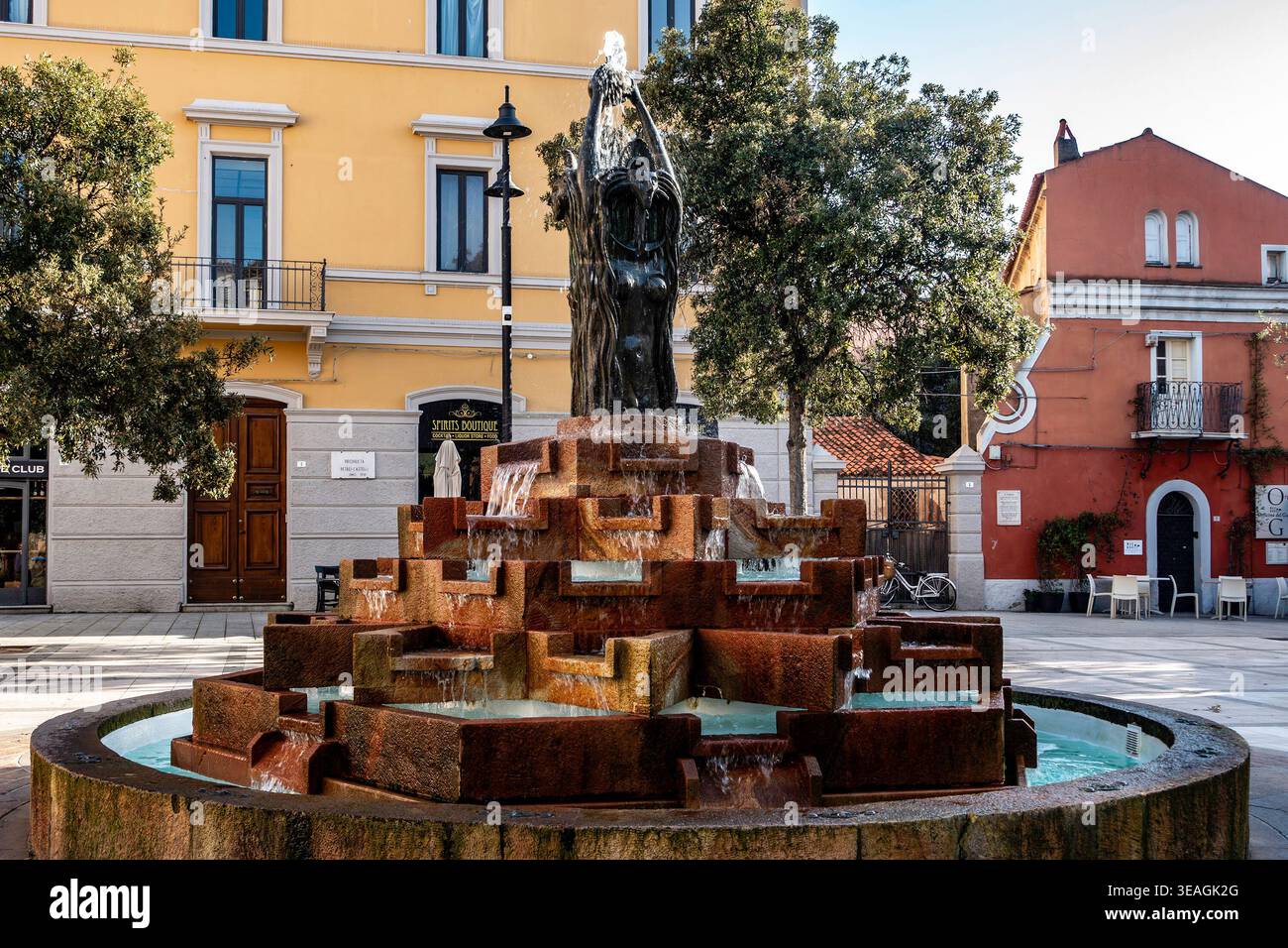 L'acqua scorre dalla fontana trivenere in pietra a gradoni di Olbia, in Sardegna, situata in una piazza lastricata circondata da edifici storici e alberi. Foto Stock
