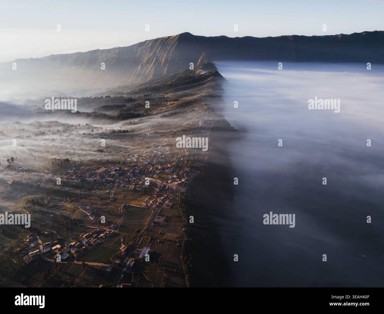 Vista aerea del Parco Nazionale di Bromo Tengger Semeru con il villaggio di Cemoro Lawang arroccato su un bordo di una scogliera sopra un mare denso di nuvole all'alba Bro Foto Stock