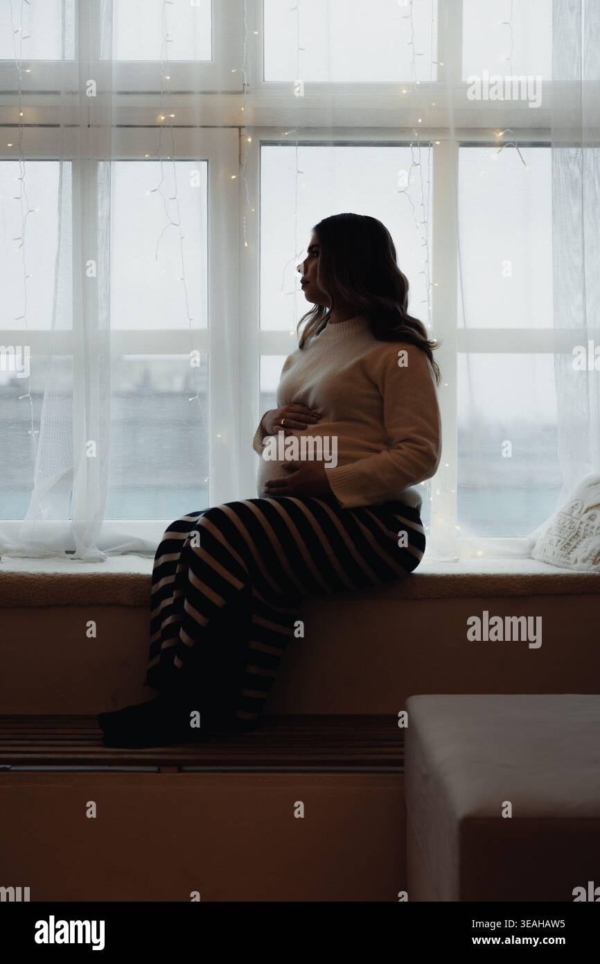 Seduta di donna silhouette con finestrino, appunti per scrittrice contemporanea, comodo pullover e gonna a righe, luce soffusa del mattino durante la pioggia Foto Stock