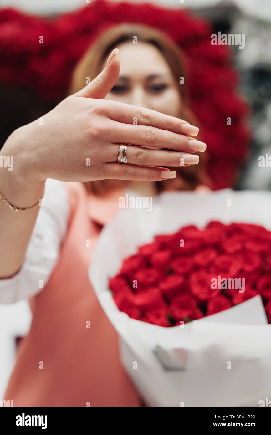 Le mani in primo piano mostrano l'anello di fidanzamento mentre la donna tiene il bouquet di rose rosse sullo sfondo, un'espressione gioiosa, colori caldi e sfondo festoso Foto Stock