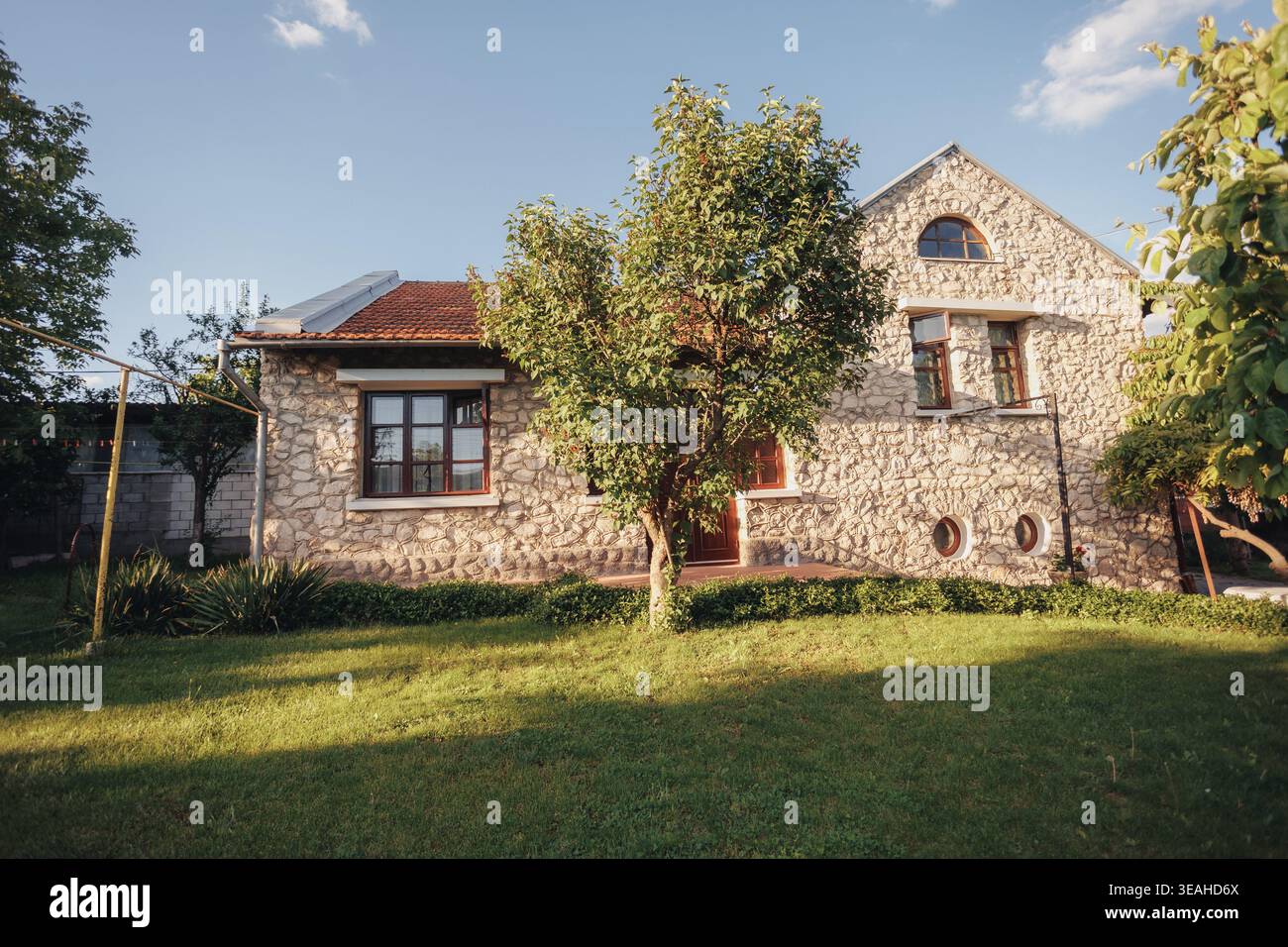 Esterno della casa in pietra con giardino illuminato dal sole, rifugio di campagna degli scrittori bagnato dalla calda facciata di pietra chiara del pomeriggio, tetto piastrellato, cornici finestre in legno Foto Stock