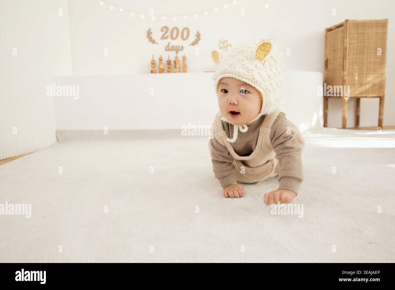 Un bambino con un cappello di pecora giace su un pavimento bianco, guardando intorno in una stanza illuminata dal sole. Foto Stock