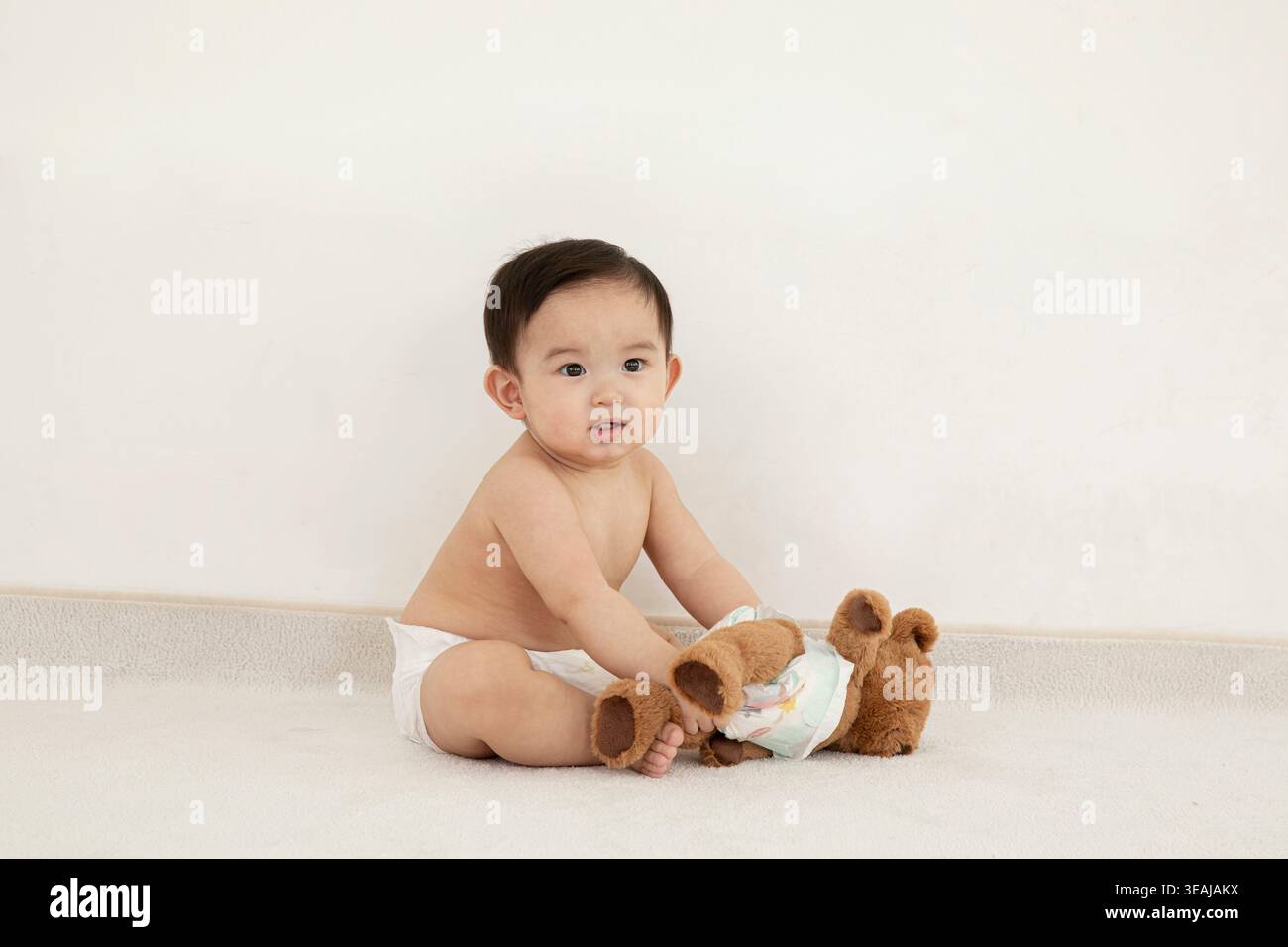 Un bambino in un pannolino siede su un pavimento bianco, tenendo un orsacchiotto marrone in un pannolino bianco. Foto Stock