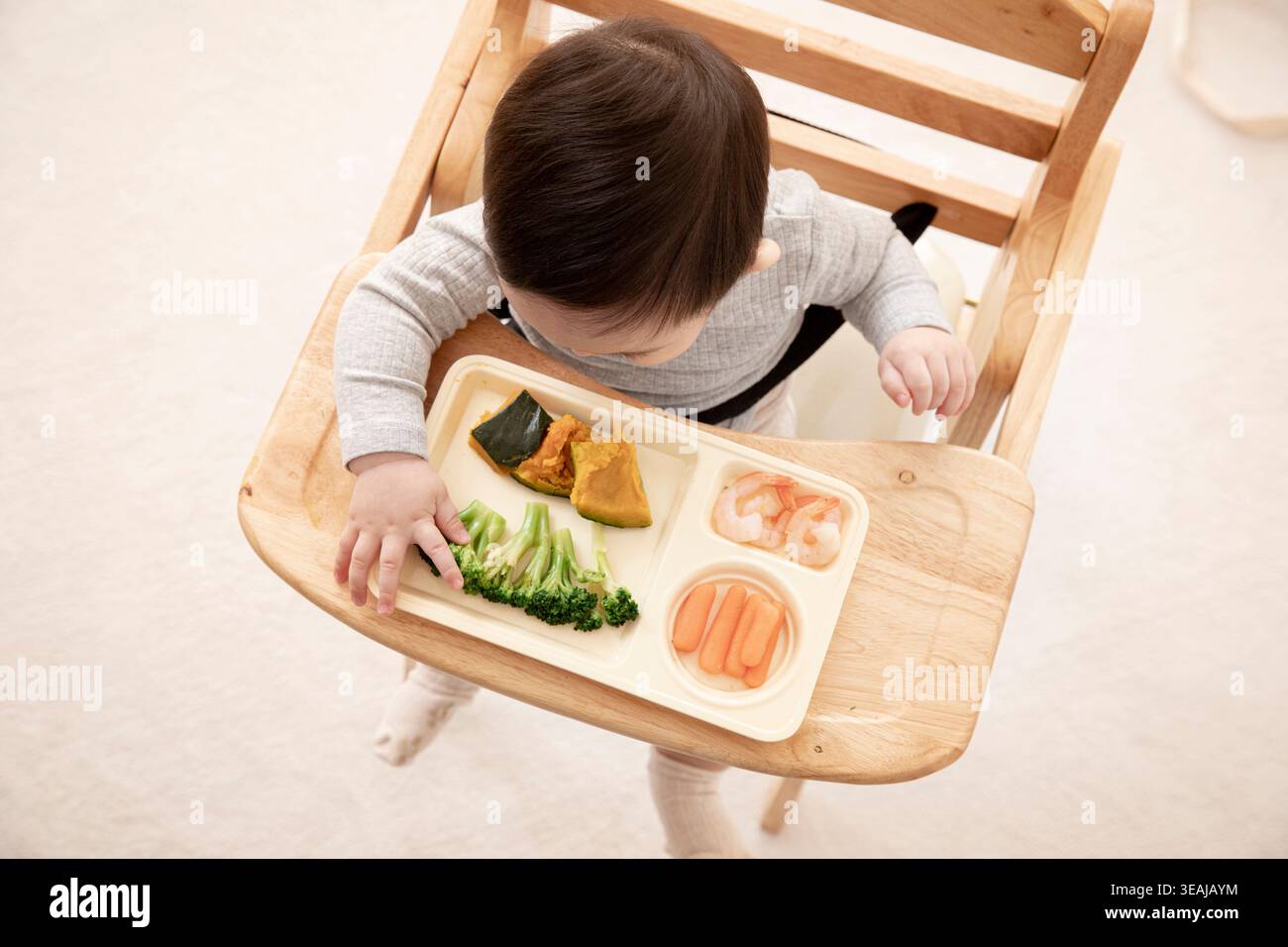 Un bambino sta mangiando un pasto a base di broccoli, carote, gamberi e zucca in un seggiolone. Foto Stock