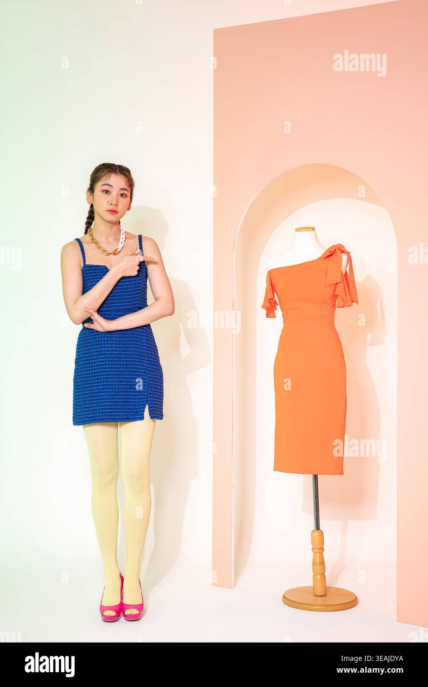 Una giovane donna con un abito blu si erge accanto a un vestito arancione su un manichino, entrambi in una stanza bianca neutra. Foto Stock