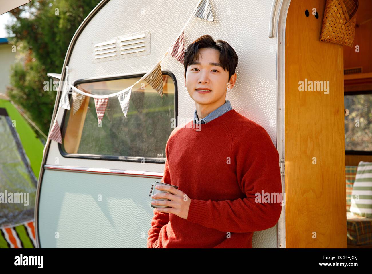 Un giovane con un maglione rosso si trova di fronte a un camper d'epoca, tenendo in mano una tazza e sorridendo davanti alla macchina fotografica. Foto Stock