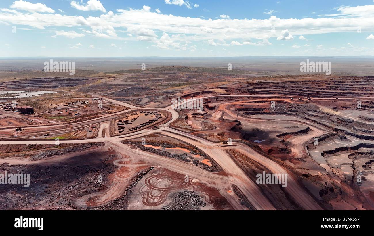 Vista aerea dell'esteso paesaggio cicatrizzato di una miniera a cielo aperto, dove la terra rossa si contrappone nettamente con il cielo azzurro pallido e l'orizzonte lontano Foto Stock