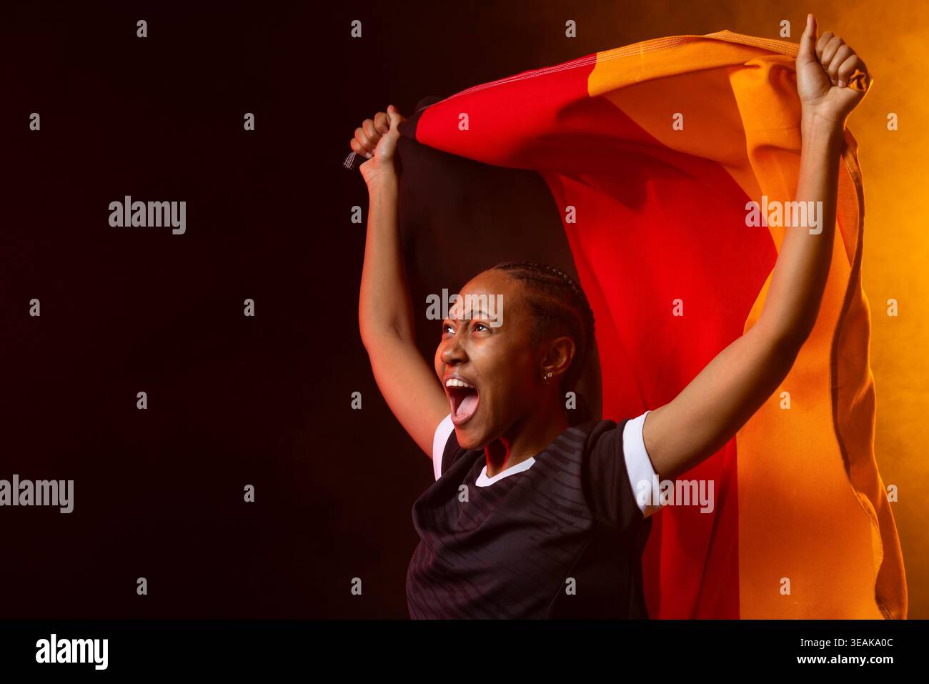 Donna africana che urla e tiene la bandiera tricolore sopra l'alto indossando una maglia sportiva nera in studio. Atleta, donna, energia, celebrazione, tessuto, stadio, Foto Stock