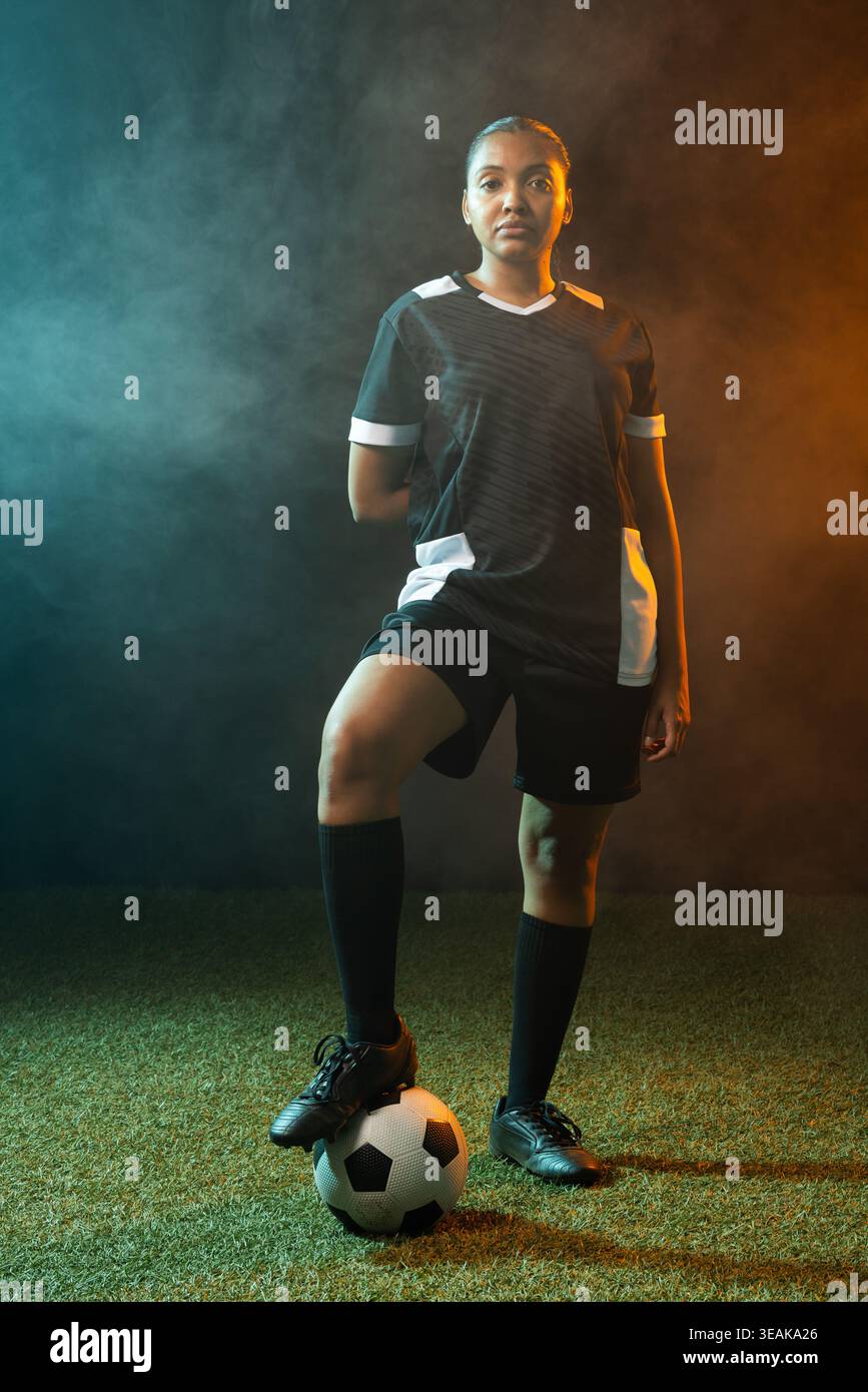 Giocatrice di calcio in piedi su un tappeto erboso con maglia nera, piede su palla in mezzo al fumo color ambra-verde. Atleta, studio, ritratto, duotono, drammatico, illuminazione Foto Stock