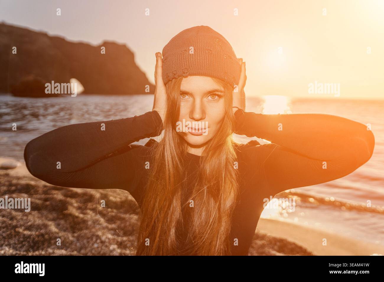 Donna, tramonto, spiaggia, che copre le orecchie con un cappello a maglia si trova vicino all'arco dell'oceano all'ora d'oro. Foto Stock