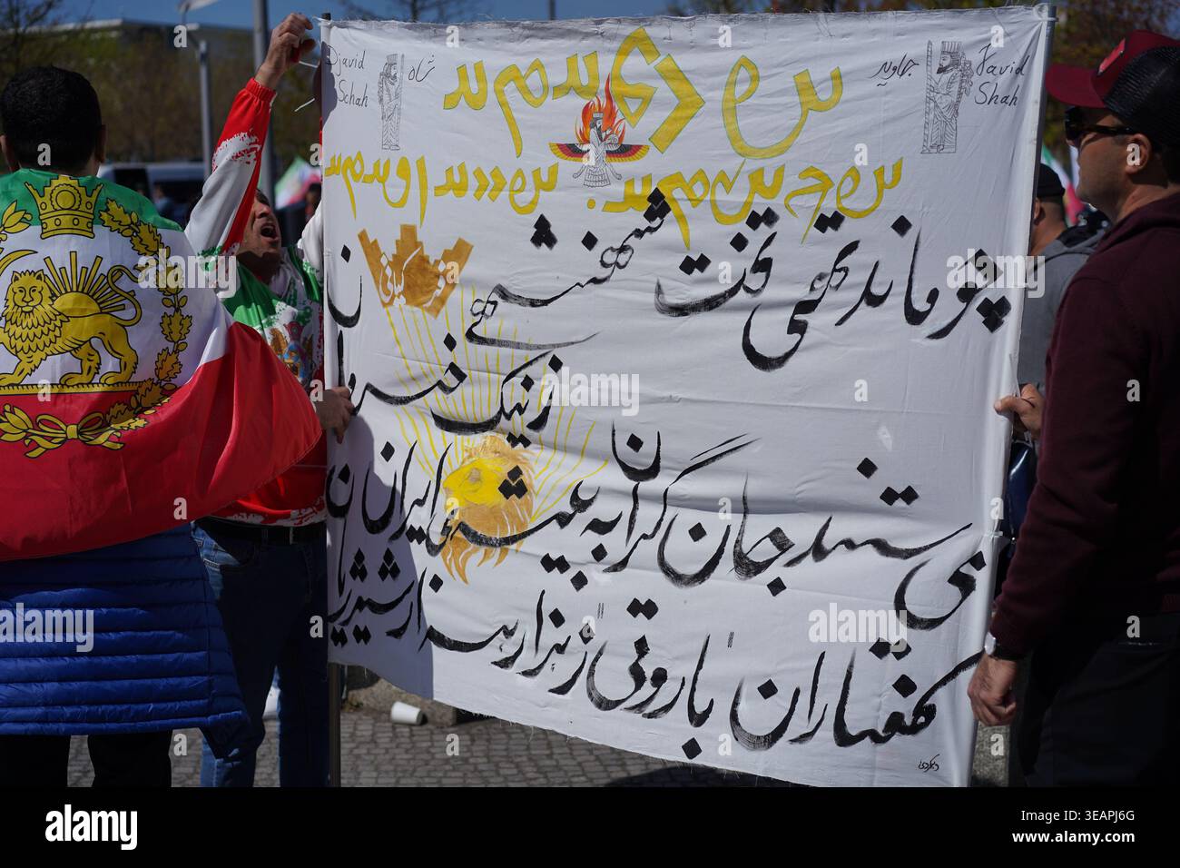 Berlino, Germania - 23 aprile 2026: I manifestanti tengono uno striscione con calligrafia persiana e simboli zoroastriani durante un raduno pro-Pahlavi a Berlino. Foto Stock
