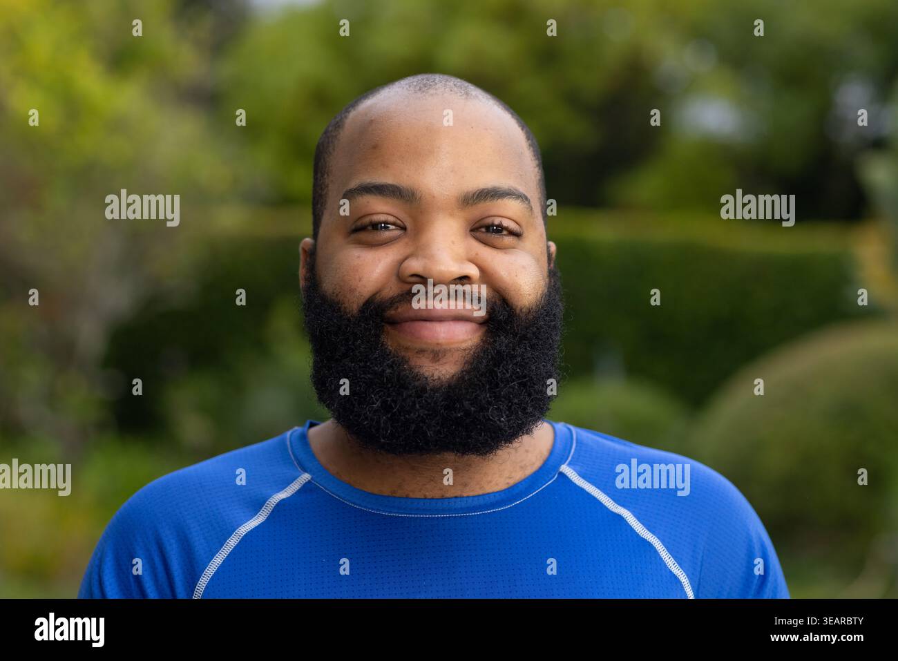 Uomo afroamericano in piedi e sorridente mentre indossa una camicia blu atletica nel giardino del cortile. Headshot, ritratto, barba, abbigliamento sportivo, outdoor, boke Foto Stock