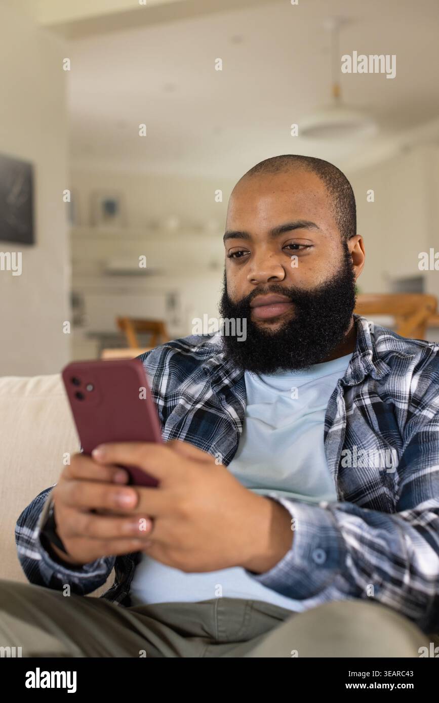 Uomo afroamericano seduto su un divano leggero a casa con un telefono color marrone, con una camicia a quadri. Casual, lifestyle, moderno, portatile, smartphone, ambiente Foto Stock