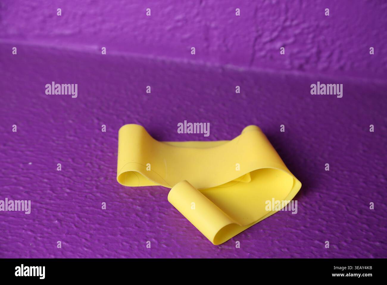 Un elastico giallo è adagiato su una parete viola. La banda ha una forma curva e mostra alcune ombre sulla superficie ruvida. La scena è luminosa e semplice. Foto Stock