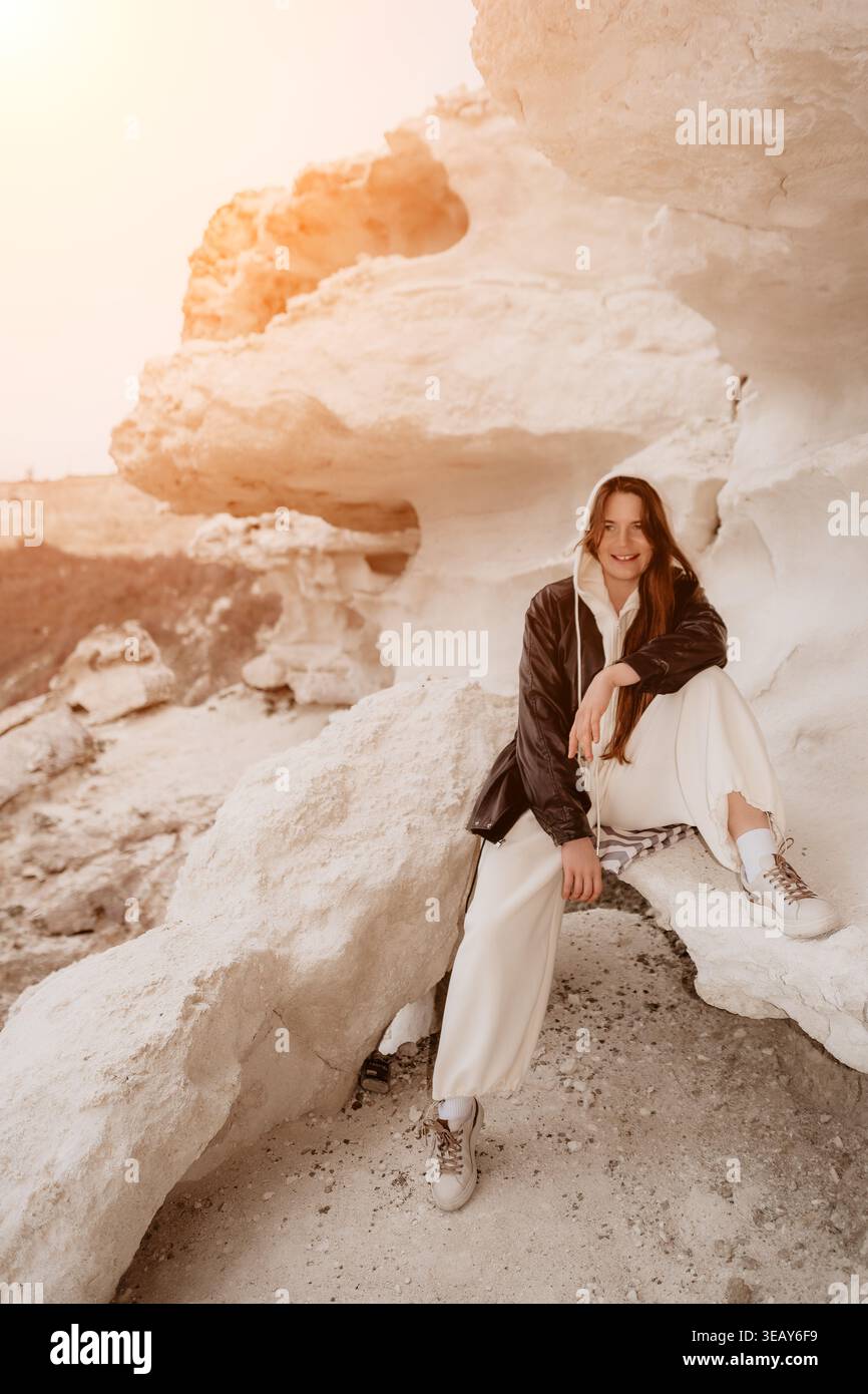 Donna in mezzo alla natura di roccia seduta su una formazione rocciosa bianca al tramonto, indossando una giacca nera e una felpa con cappuccio Foto Stock